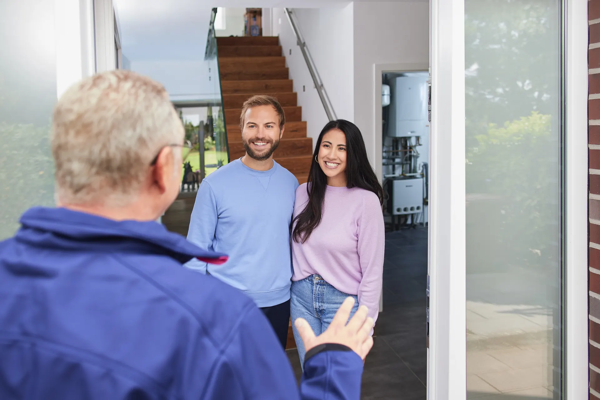 Ein Handwerker begrüßt eine junges Paar freundlich in ihrem Eigenheim. Im Hintergrund ist eine geöffnete Tür mit einer modernen Heizungsanlage zu sehen. 