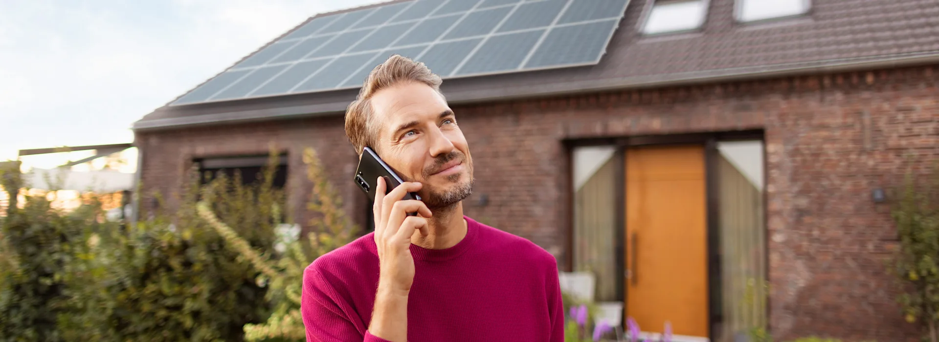 Mann telefoniert vor Photovoltaik-Anlage Ein Mann im roten Pullover steht vor seinem Einfamilienhaus auf dessen Dach eine Solaranlage installiert ist und schaut telefonierend in die Ferne.