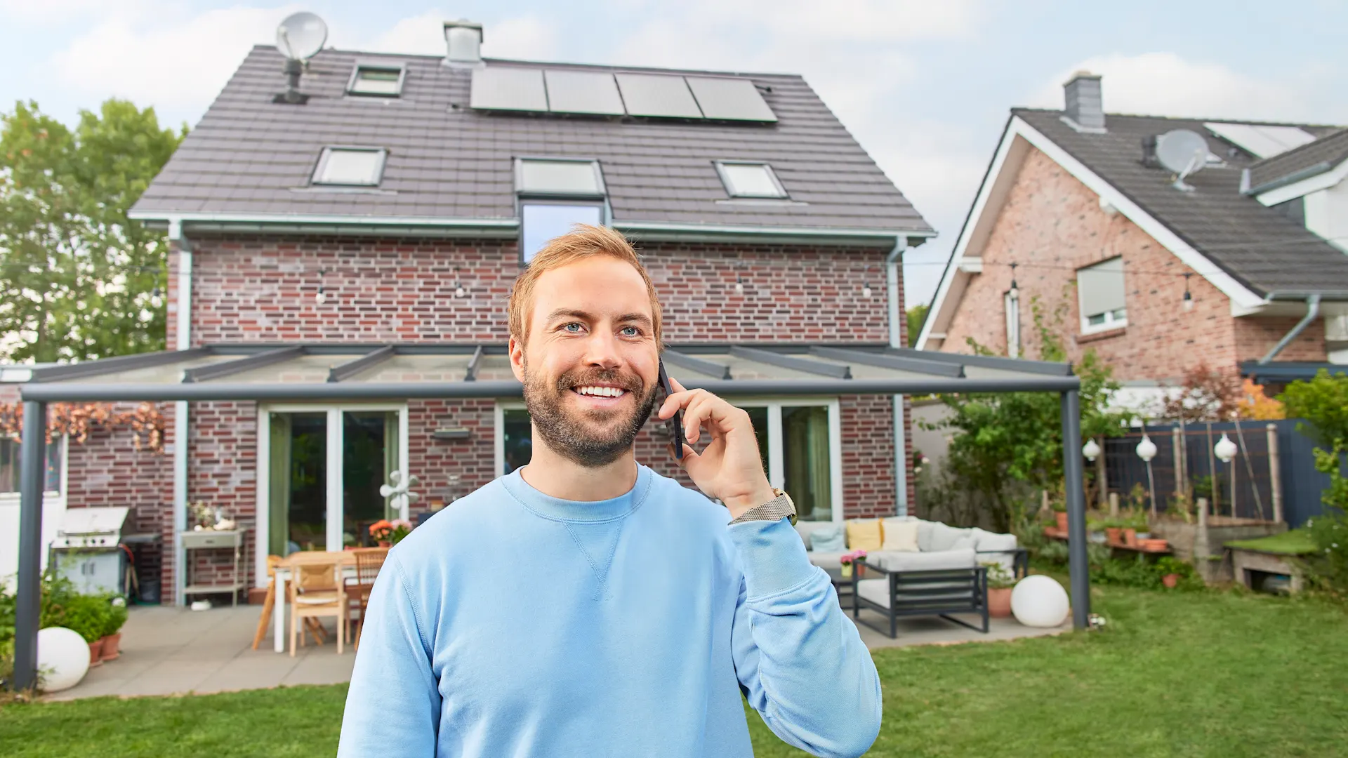 Mann telefoniert vor seinem Einfamilienhaus Zu sehen ist ein Einfamilienhaus, das aus der Perspektive des Gartens fotografiert ist. Der Mann steht im Vordergrund und telefoniert mit einem Smartphone.