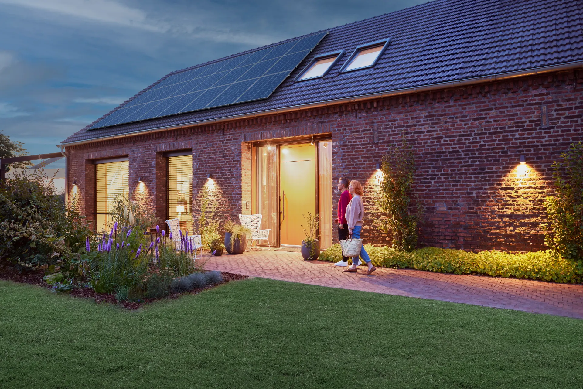 Ein Einfamilienhaus auf dessen Dach eine Photovoltaikanlage zu sehen ist. Es ist Abenddämmerung und das Haus scheint hell erleuchtet. 