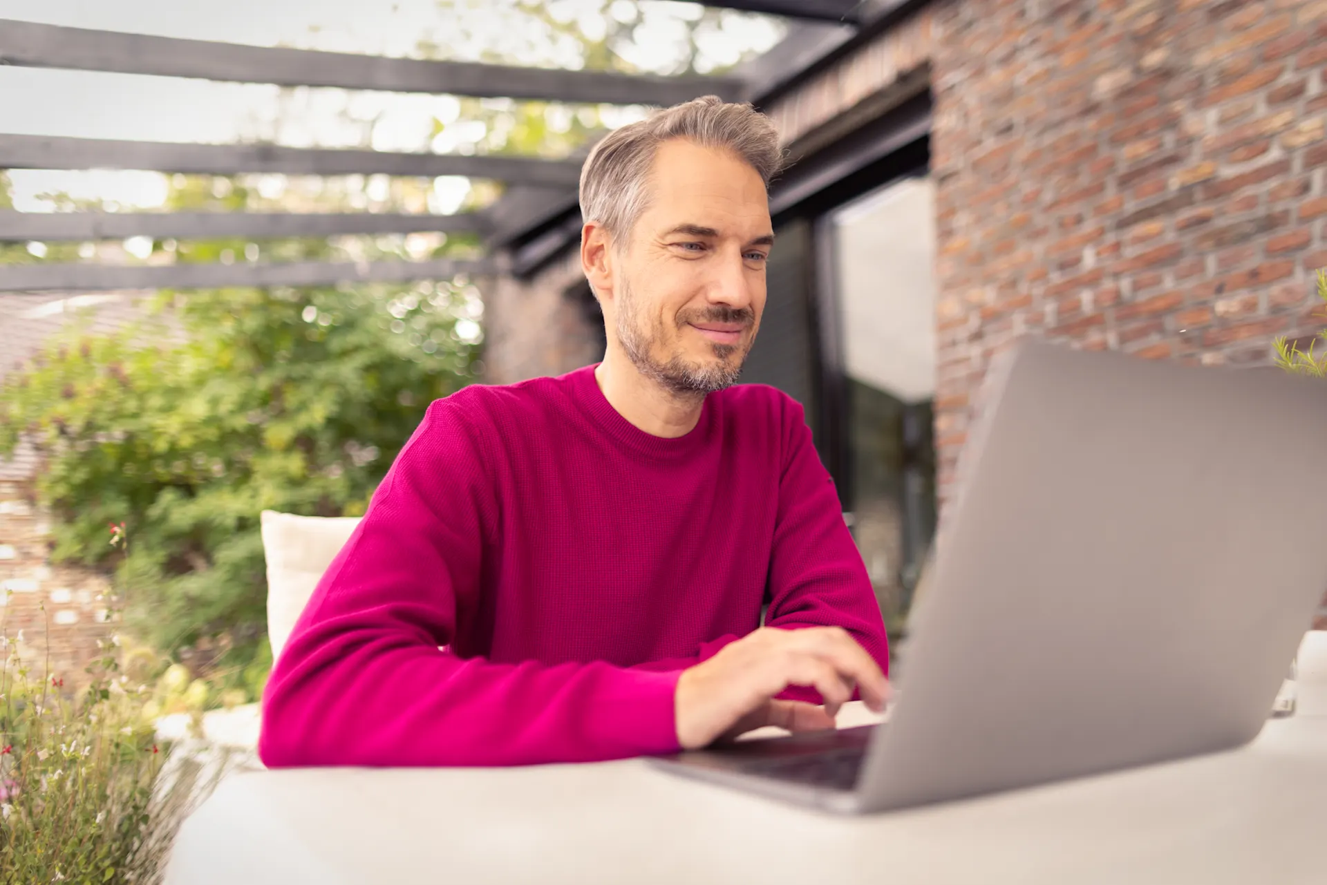 Ein Mann mit graumelierten Haaren und einem pinken Pullover sitzt entspannt an einem Tisch im Freien und arbeitet lächelnd an seinem Laptop.