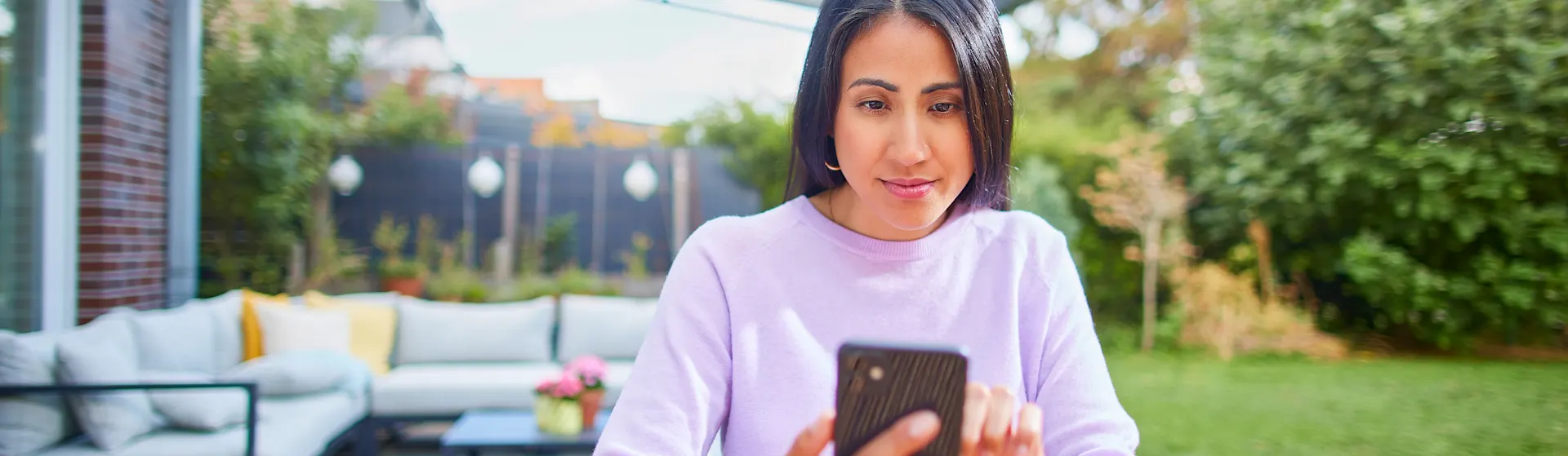 NEW Dienstleistungen Eine Frau sitzt im Garten mit ihrem Smartphone und informiert sich über die Dienstleistungen und den Service bei der NEW Energie.