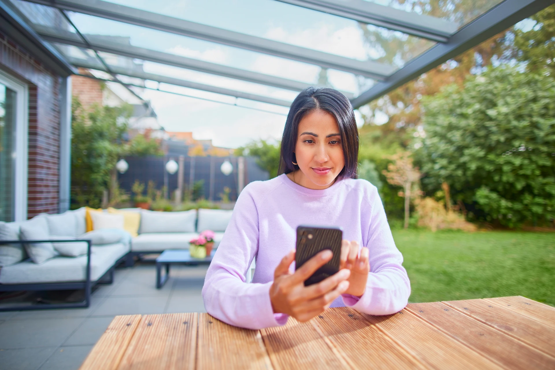 Eine Frau sitzt im Garten mit ihrem Smartphone und informiert sich über die Dienstleistungen und den Service bei der NEW Energie.