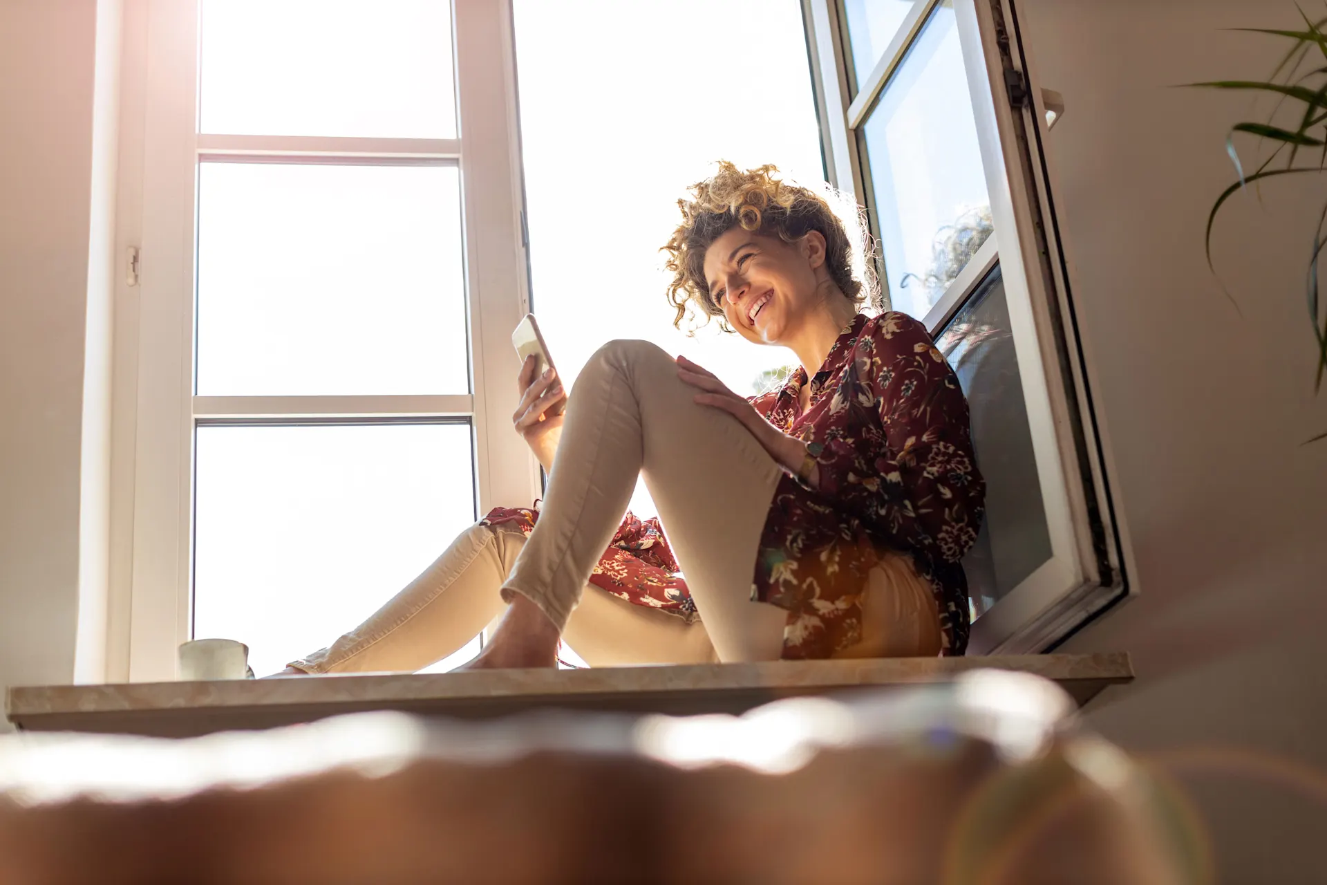 Frau mit einem Smartphone in der Hand sitzt auf einer Fensterbank vor dem geöffneten Fenster.