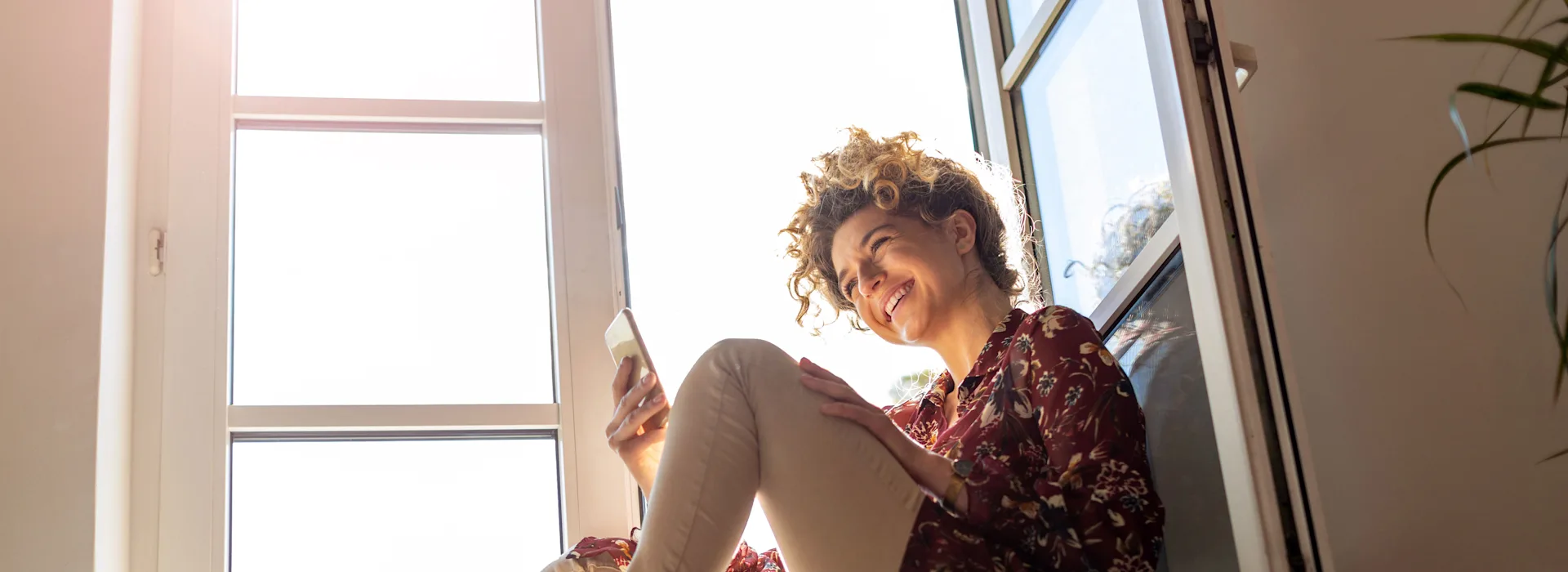 Attraktive Tarife bei der NEW Energie Frau mit einem Smartphone in der Hand sitzt auf einer Fensterbank vor dem geöffneten Fenster.