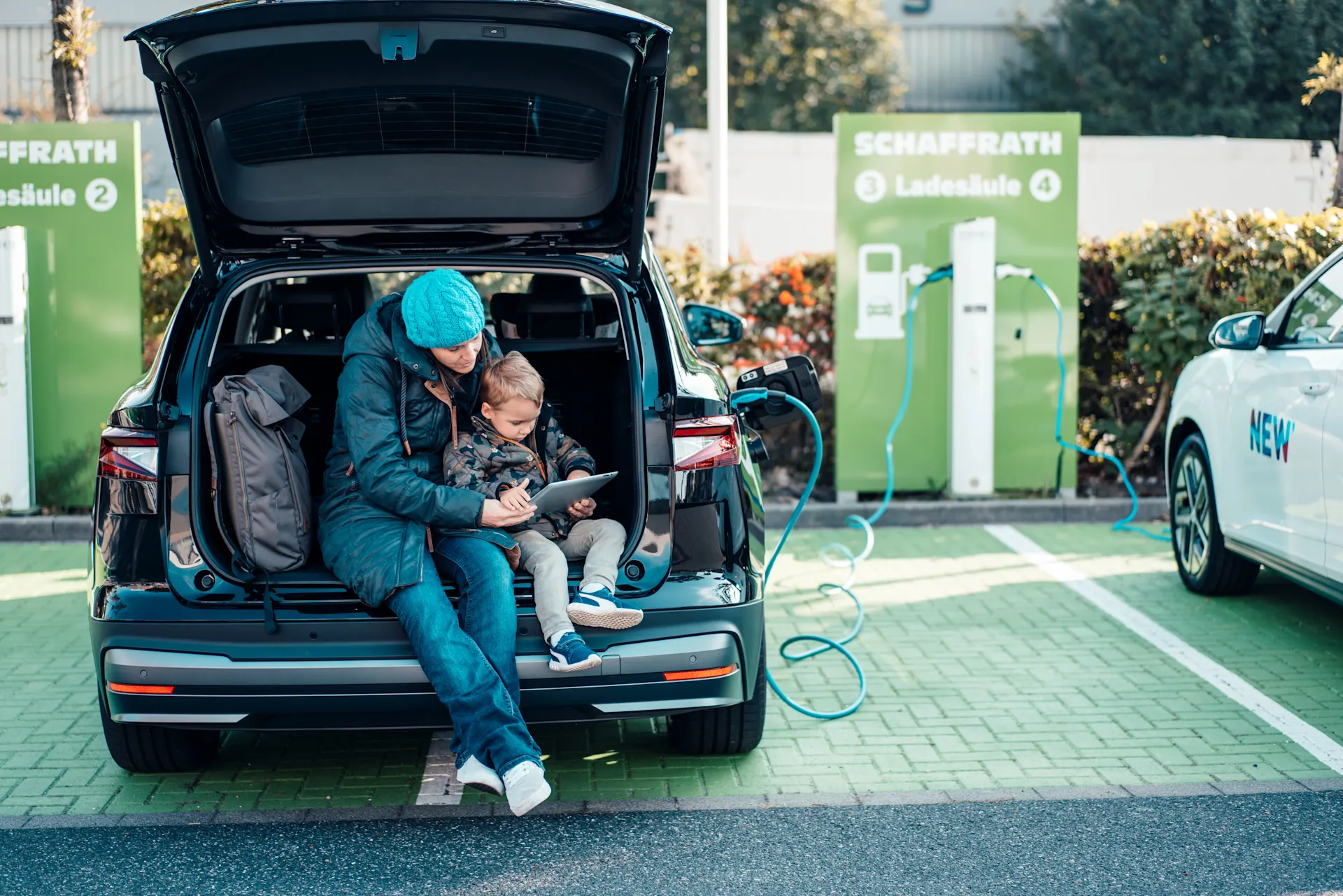 Mutter und Sohn sitzen im Kofferraum ihres E-Autos und verfolgen am Tablet den Ladevorgang ihres Autos. Dies steht an einer Ladesäule der NEW Energie.