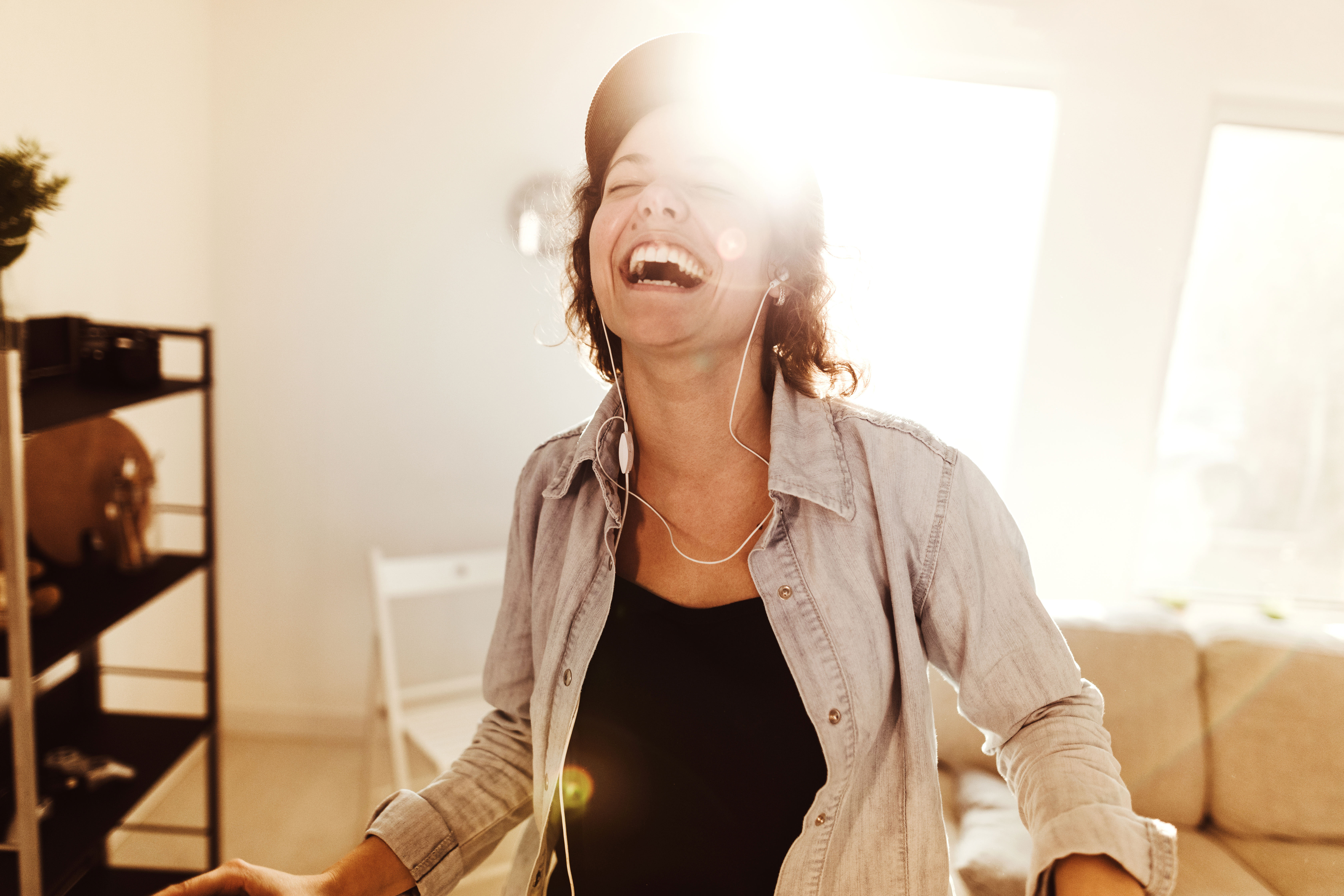 Frau steht lachend im Wohnzimmer und hört Musik über Kopfhörer. Sie freut sich über die Angebote der NEW Energie.