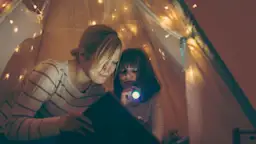 Was Sie bei einem Stromausfall tun können Mutter sitzt mit ihrer Tochter in einem beleuchteten Zelt und hat eine Taschenlampe in der Hand. Gemeinsam lesen sie ein Buch.