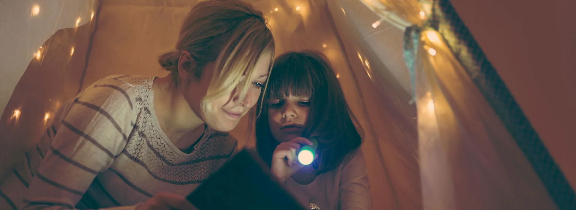Was Sie bei einem Stromausfall tun können Mutter sitzt mit ihrer Tochter in einem beleuchteten Zelt und hat eine Taschenlampe in der Hand. Gemeinsam lesen sie ein Buch.