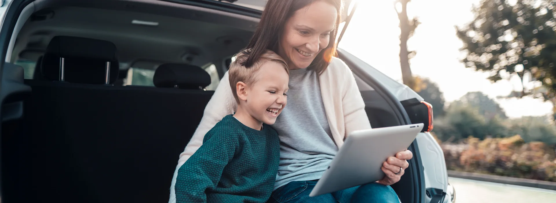 Familie informiert sich über Produkte Eine Mutter mit Kind parkt auf einem öffentlichen Parkplatz, sitzt im Kofferraum und informiert sich mit einem Tablet über den Energieversorger NEW Energie.