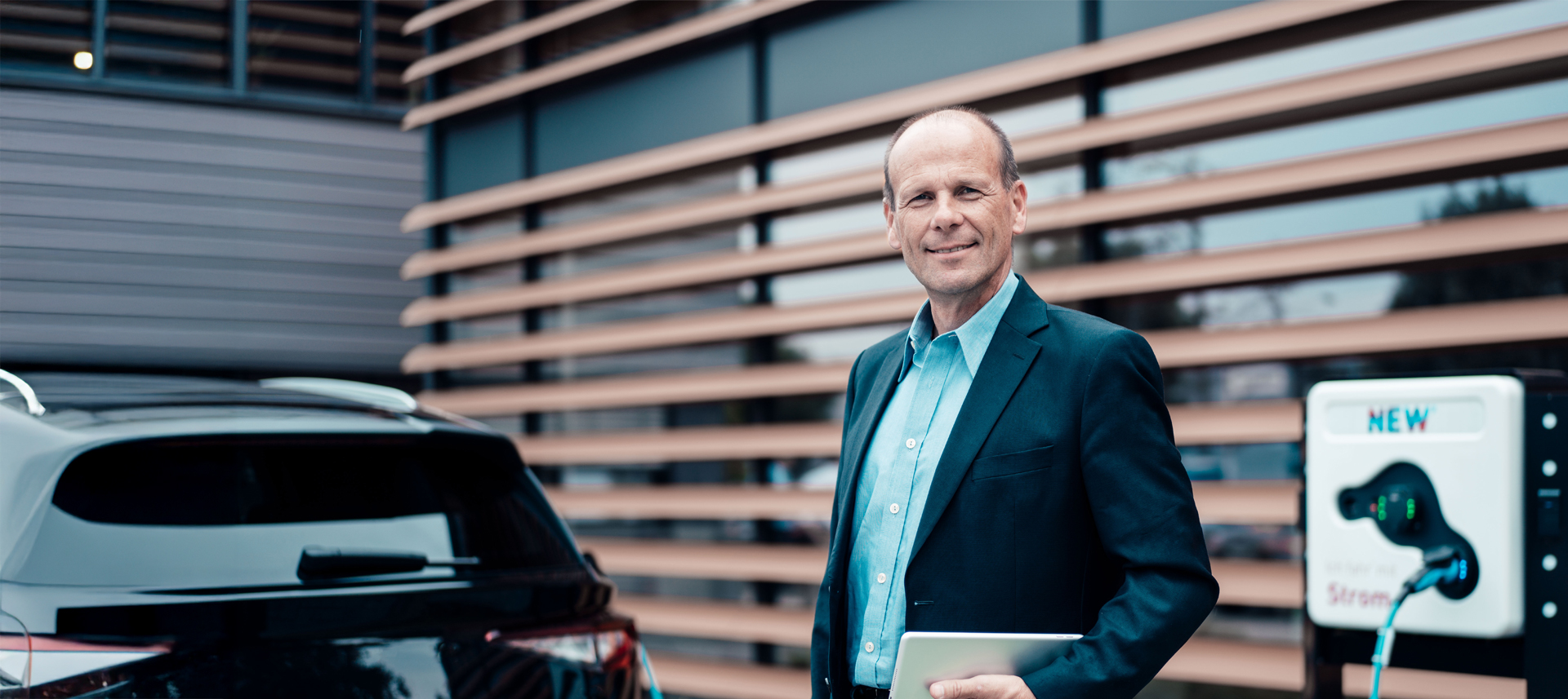 Mann steht vor Ladesäule auf einem Firmengelände und lädt sein Auto. Er lächelt in die Kamera.