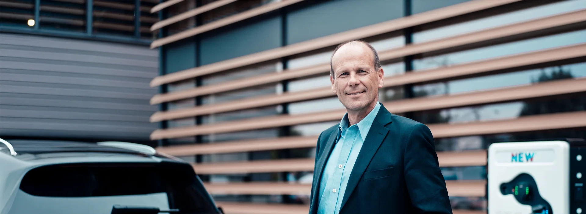 Ladelösungen für Ihr Unternehmen Mann steht vor Ladesäule auf einem Firmengelände und lädt sein Auto. Er lächelt in die Kamera.