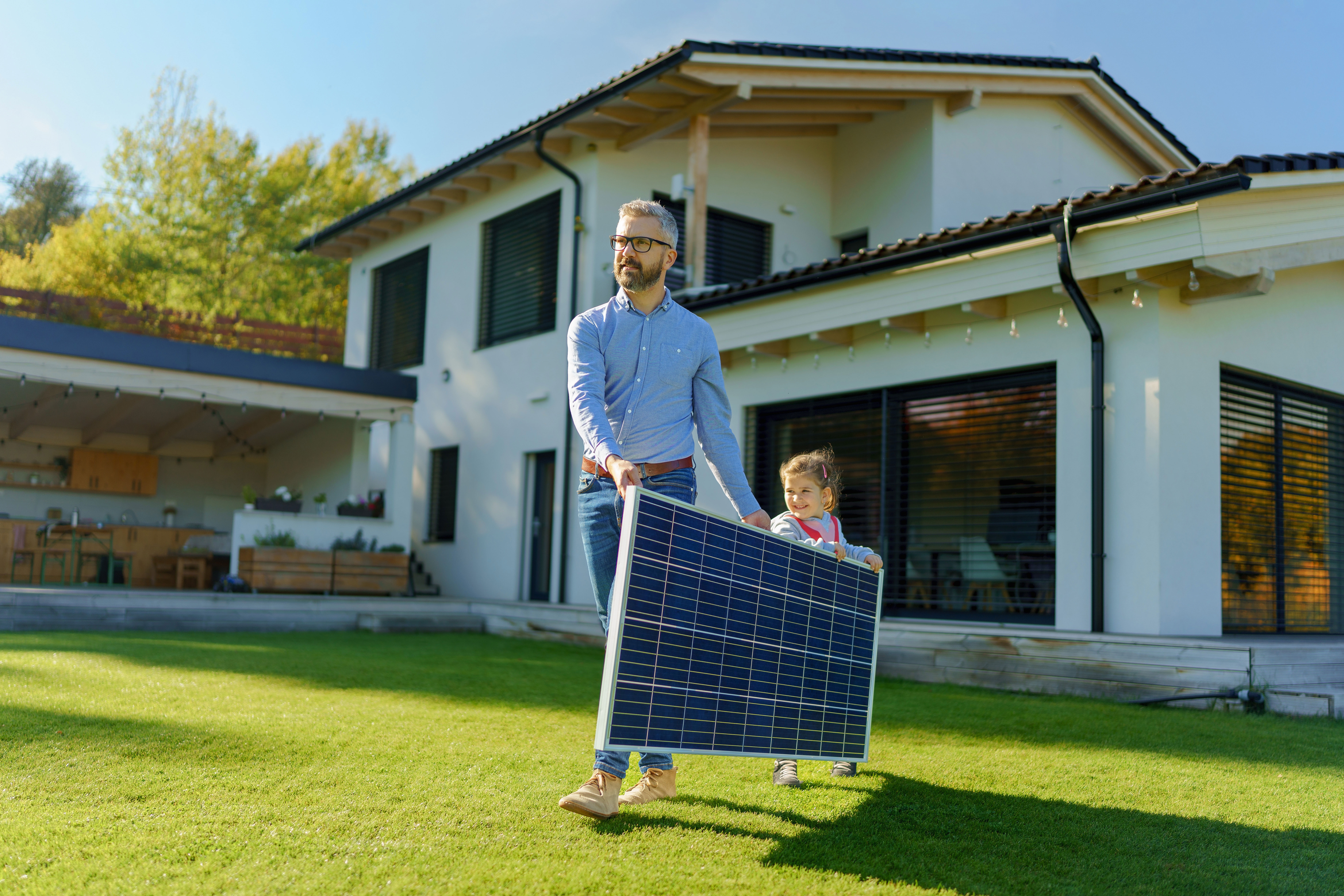 Vater und Tochter tragen ein PV Modul der NEW Energie durch den Garten ihres Einfamilienhauses.