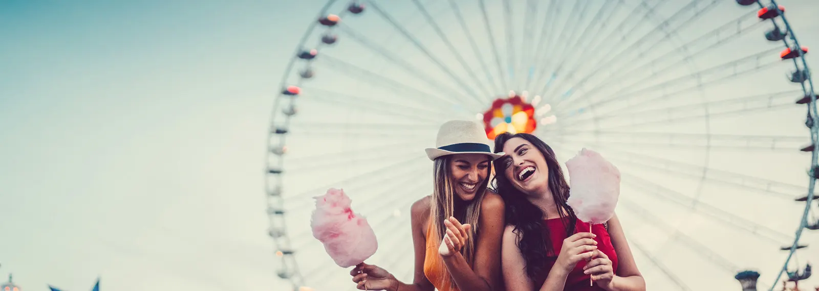 Zwei Freundinnen stehen vor einem Riesenrad und essen Zuckerwatte gemeinsam. Sie freuen sich über die Vorteile bei der NEW Energie.