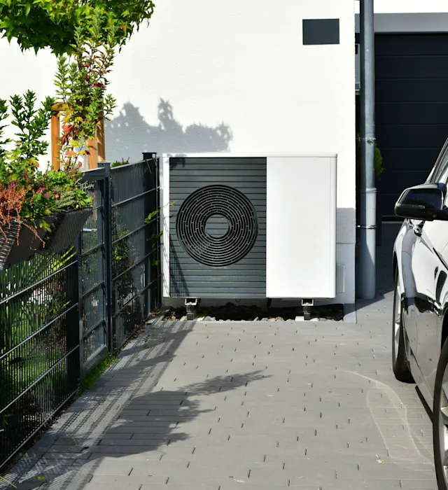 Eine Wärmepumpe steht an einer Garage im Außenbereich.