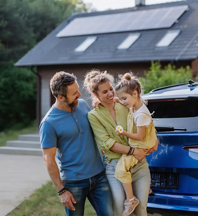 Familie vor Haus mit PV-Anlage und Elektroauto Eine Familie steht an einem Elektroauto was gerade geladen wird. Im Hintergrund ist ein Haus mit einer Photovoltaik-Anlage auf dem Dach zu sehen.