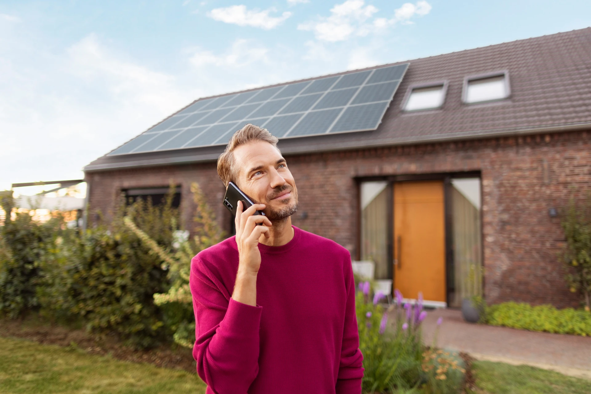 Ein Mann hat ein Telefonat im Garten vor einem Haus mit einer Photovoltaik Anlage, welche für die Erzeugung von Ökostrom genutzt wird.