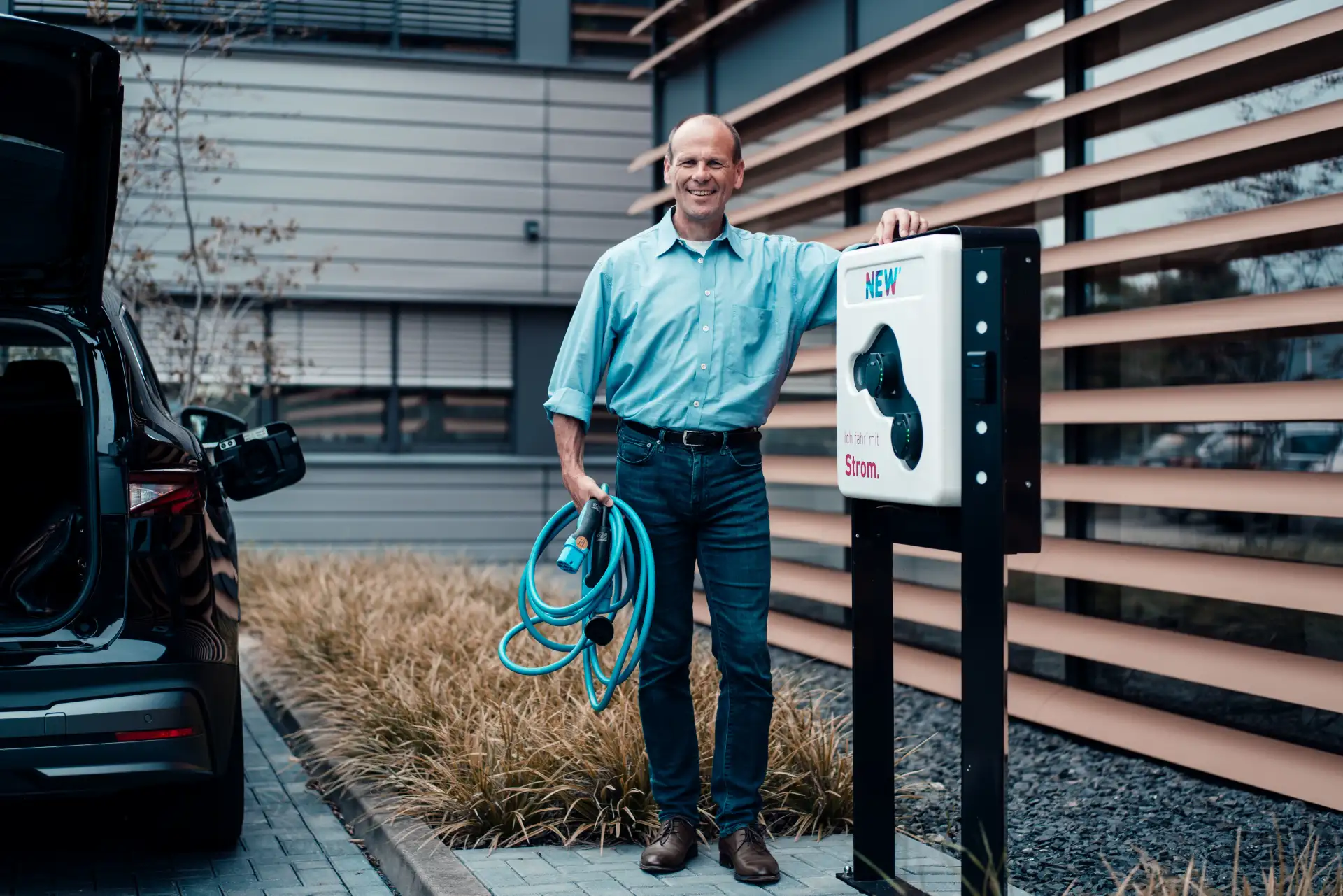 Ein Mann steht lächeln an einer Wallbox und hält ein Ladekabel in der Hand.