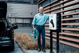 Mann angelehnt an einer Wallbox Ein Mann steht lächelnd neben einem Elektroauto an einer Wallbox und hält ein Ladekabel in der Hand.