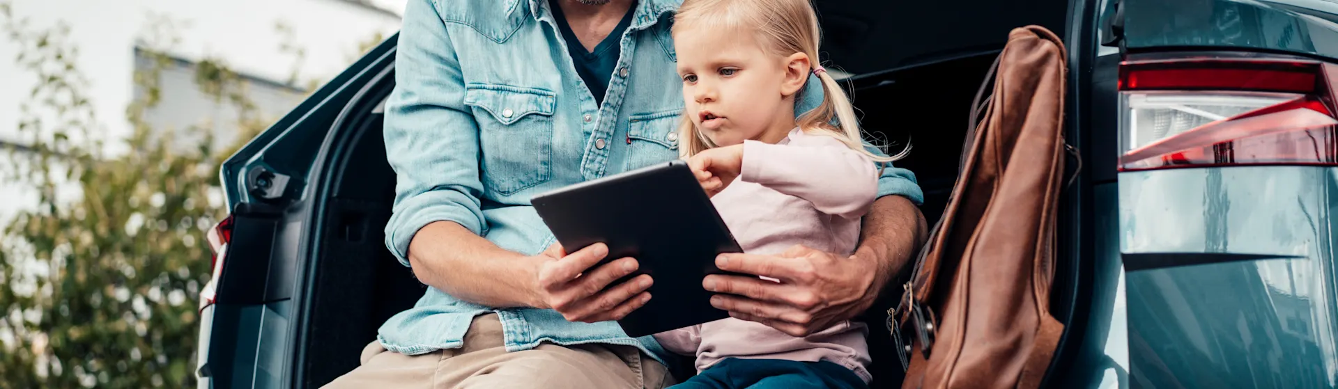 Autostromtarife vergleichen Vater und Tochter sitzen im offenen Kofferraum und vergleichen die Autostromtarife der NEW Energie über ein Tablet.