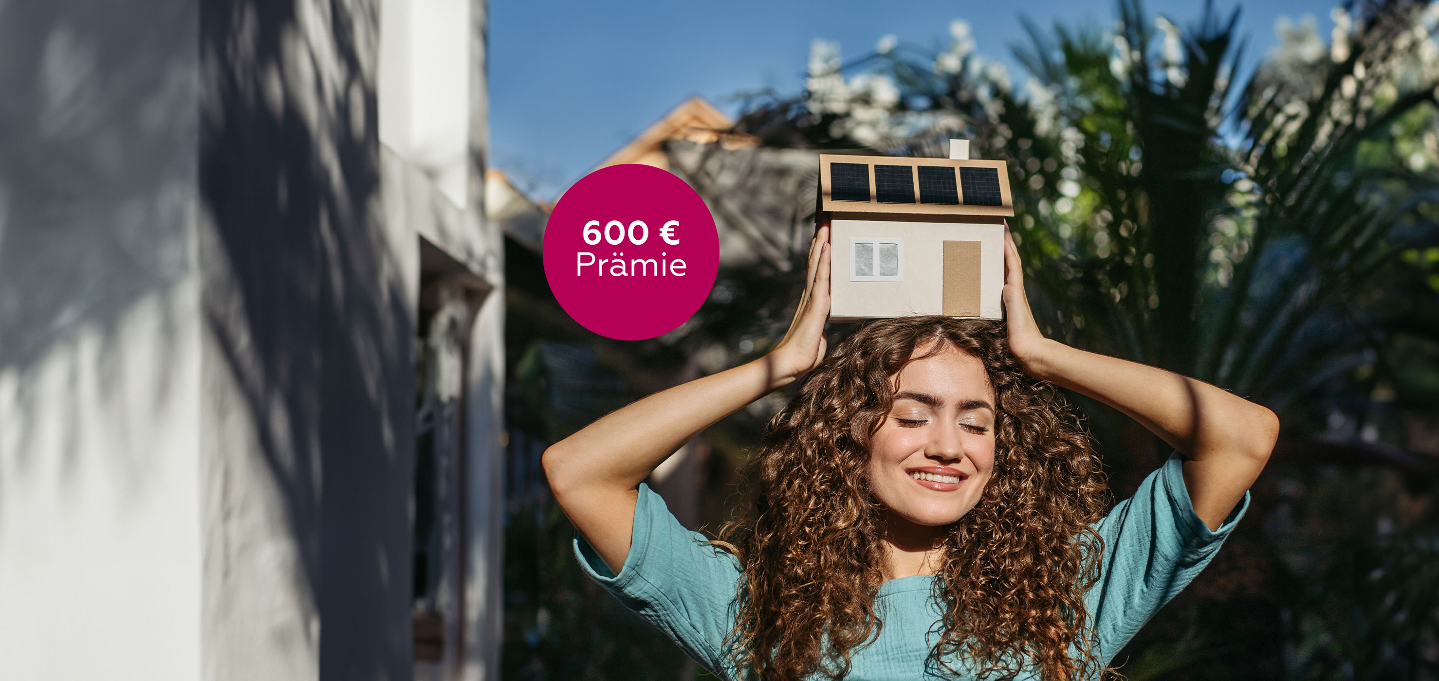 Frau hält sich draußen ein kleines Haus-Modell mit Solarpaneelen auf dem Dach über den Kopf.