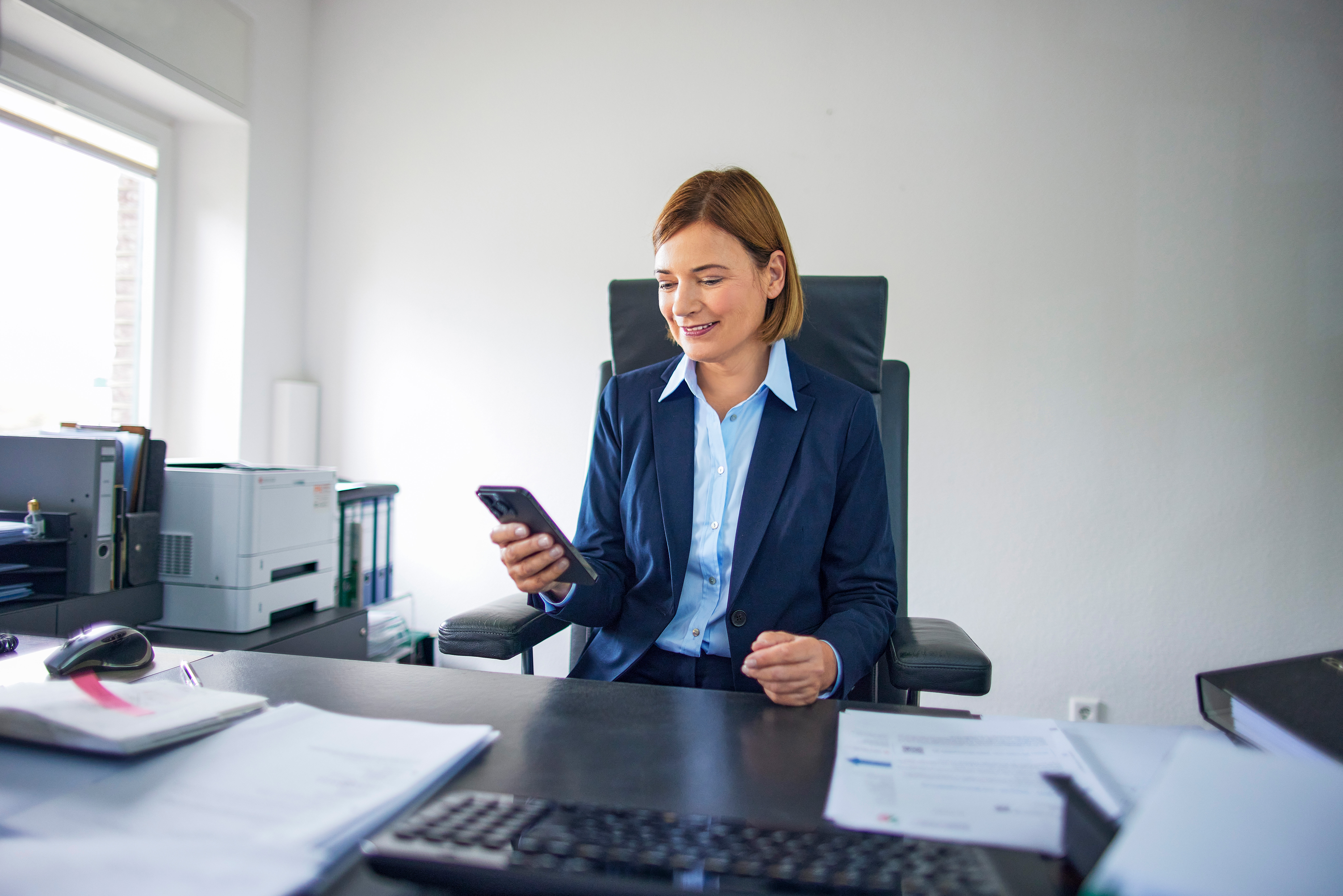 Eine Frau im Hosenanzug sitzt in einem Büro und schaut auf ihr Smartphone.