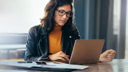 Frau macht sich mit NEW Energie vertraut Eine Frau mit schwarzer Brille sitzt am Tisch vor ihrem Laptop und schaut sich die NEW Energie an.