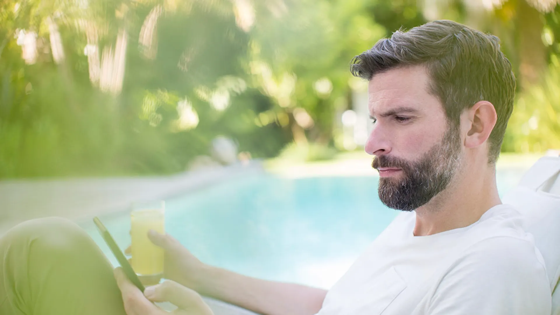 Mann an Pool Ein Mann sitzt an einem Pool mit seinem Handy und einem Getränk in der Hand. Dabei schaut er nach Stromtarifen der NEW Energie.