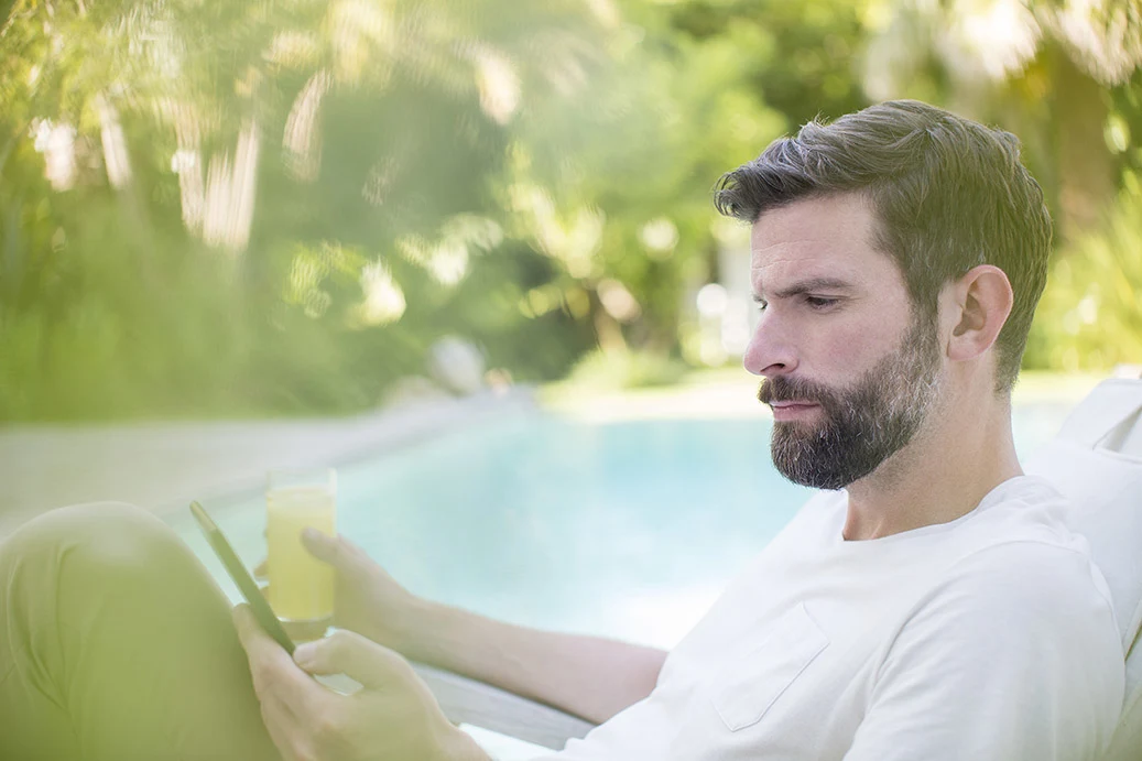 Ein Mann sitzt an einem Pool mit seinem Handy und einem Getränk in der Hand.