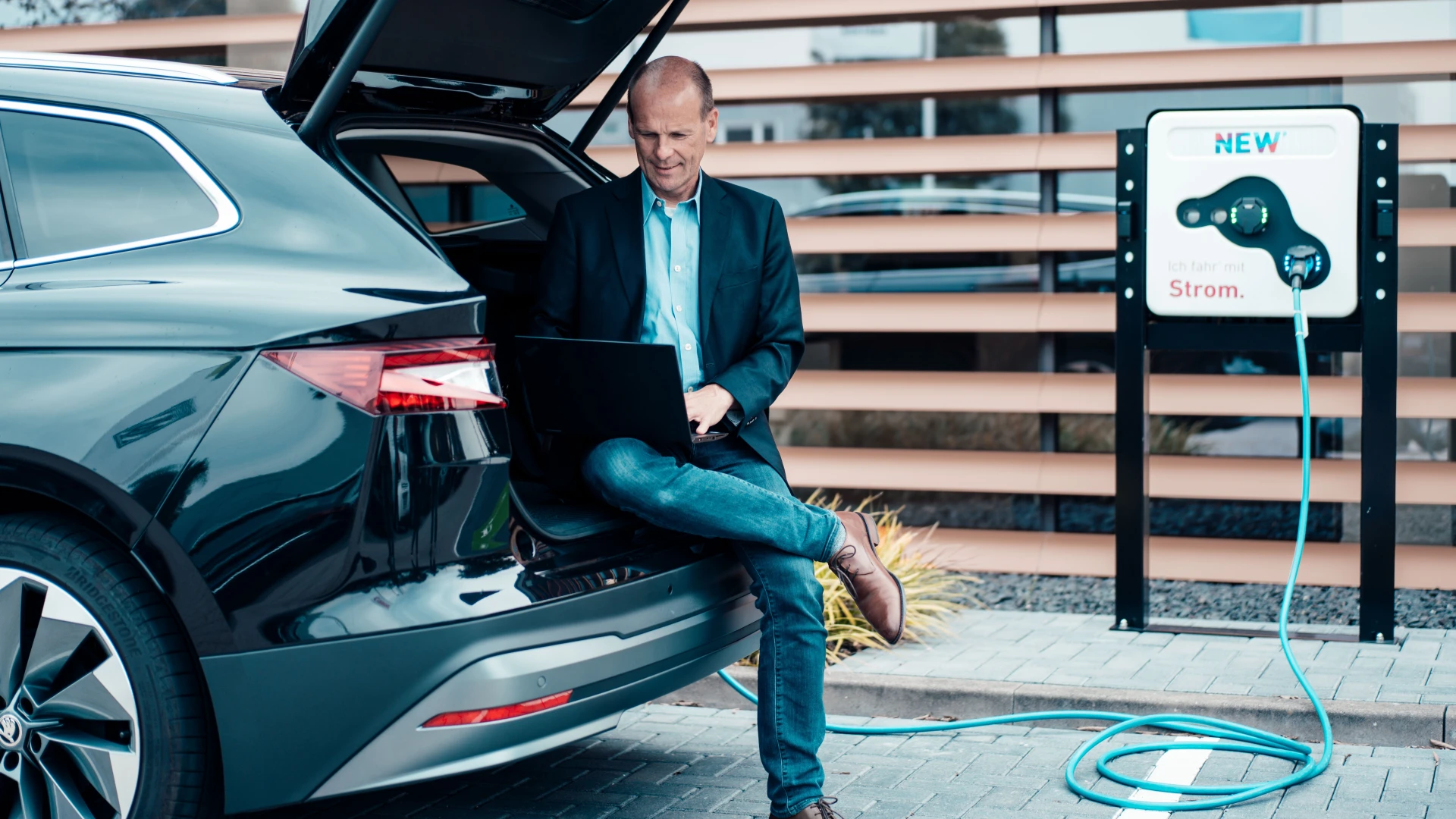 Ein Unternehmer sitzt in dem Kofferraum eines E-Autos mit seinem Laptop und schaut nach dem Service der NEW Energie.