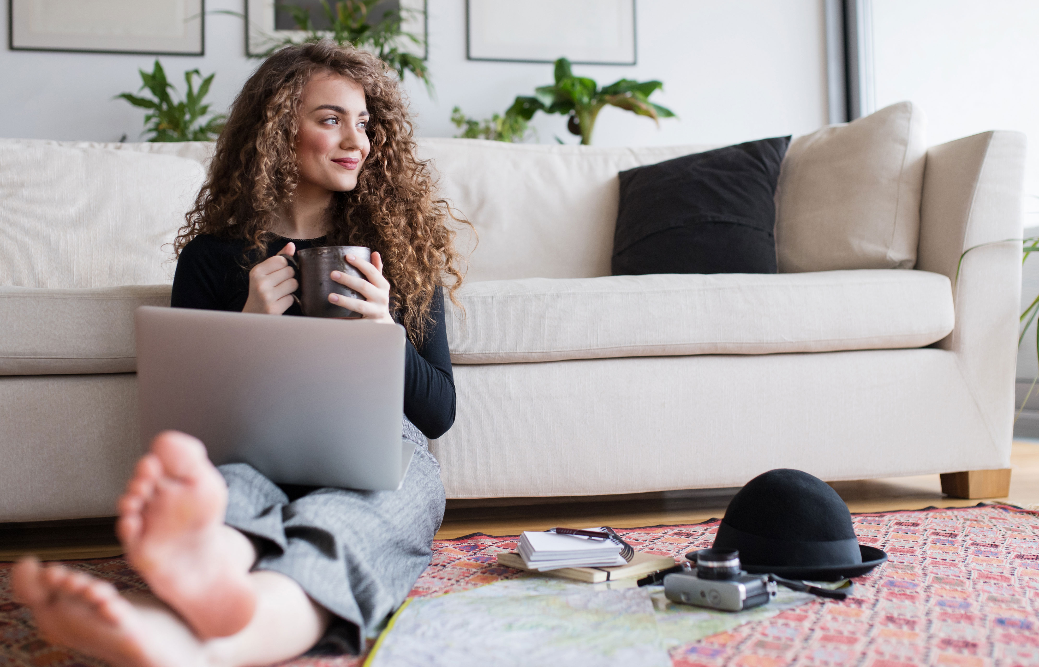 Frau mit Laptop und Kaffee sitzt entspannt vor dem Sofa auf dem Boden und schaut Tarife der NEW-Energie.