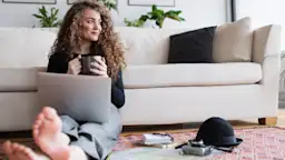 Frau mit Laptop und Kaffee Frau mit Laptop und Kaffee sitzt entspannt vor dem Sofa auf dem Boden und schaut Tarife der NEW-Energie.