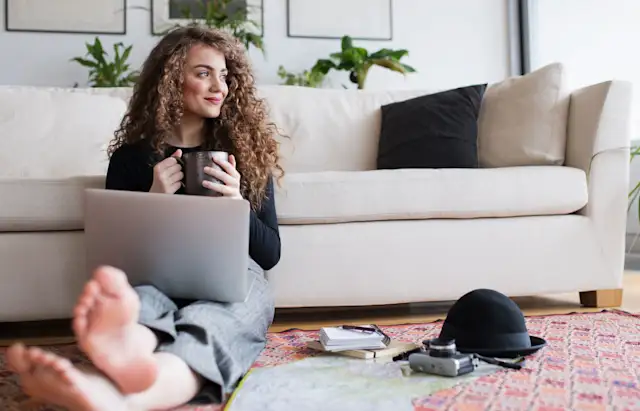 Frau mit Laptop und Kaffee Frau mit Laptop und Kaffee sitzt entspannt vor dem Sofa auf dem Boden und schaut Tarife der NEW-Energie.