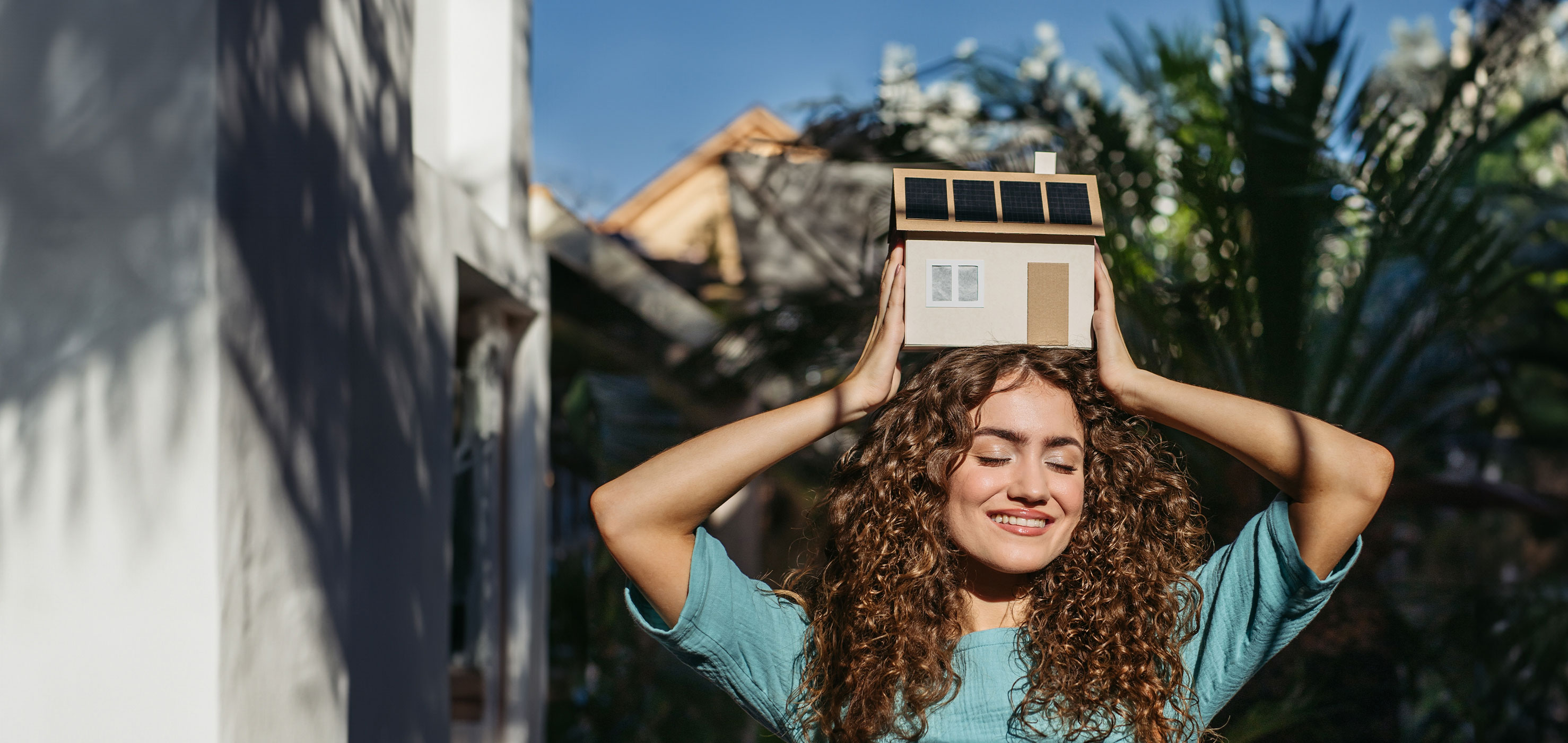 Eine Frau hält über ihrem Kopf ein Hausmodell, was eine PV-Anlage der NEW-Energie auf dem Dach hat.