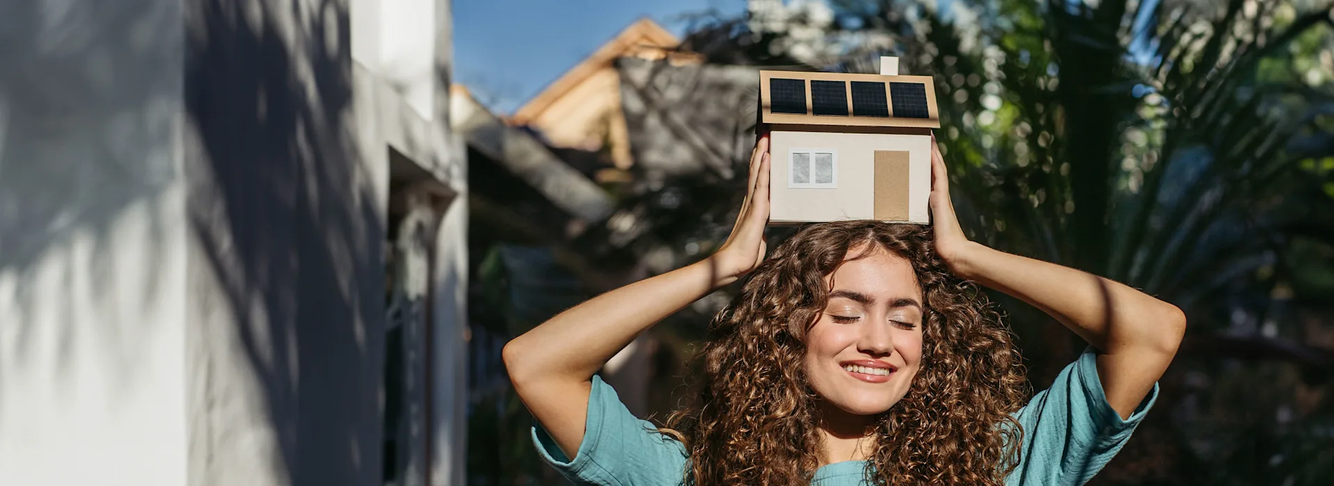Frau zeigt Hausmodell mit einer PV-Anlage Eine Frau hält über ihrem Kopf ein Hausmodell, was eine PV-Anlage der NEW-Energie auf dem Dach hat.