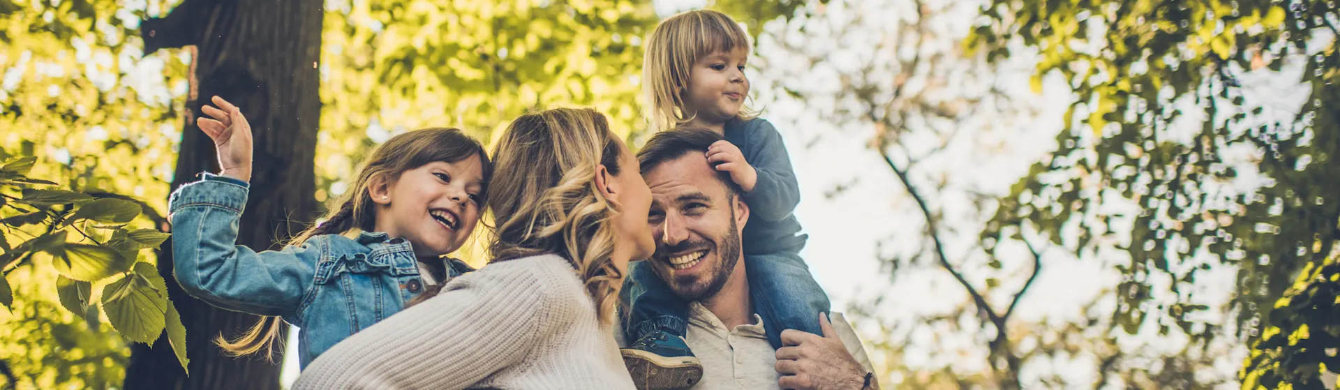 Auswahl von günstigem Ökostrom Eine Familie mit zwei Kindern ist sehr naturverbunden und befindet sich im Wald. Vater und Mutter tragen ihre Kinder auf dem Rücken. Sie nutzen Ökostrom der NEW Energie.