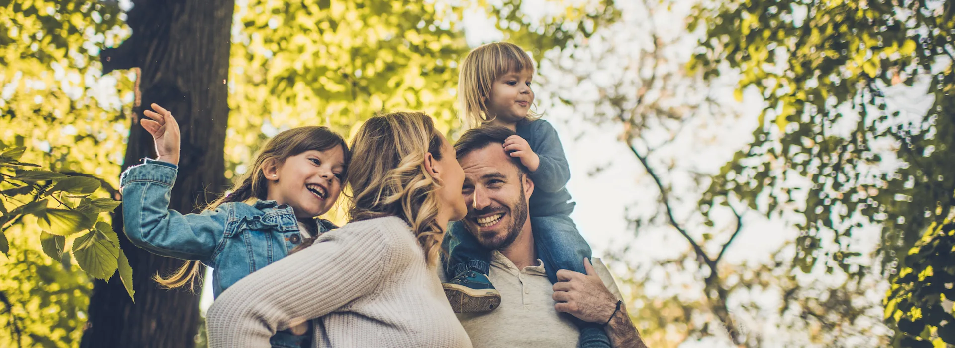 Auswahl von günstigem Ökostrom Eine Familie mit zwei Kindern ist sehr naturverbunden und befindet sich im Wald. Vater und Mutter tragen ihre Kinder auf dem Rücken. Sie nutzen Ökostrom der NEW Energie.