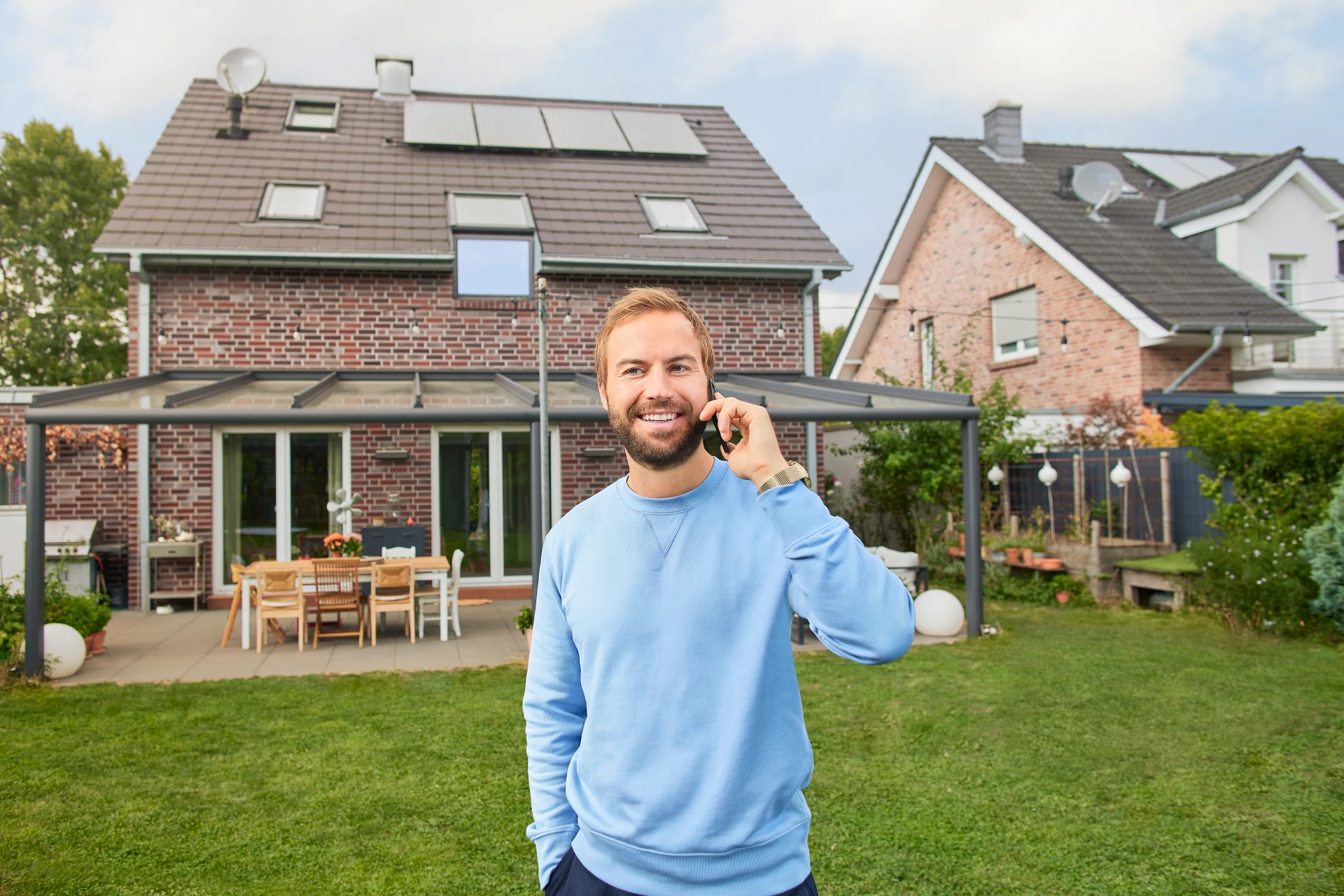 Ein Mann telefoniert mit seinem Handy im Garten vor seinem Einfamilienhaus, auf dessen Dach eine Photovoltaikanlage angebracht ist.