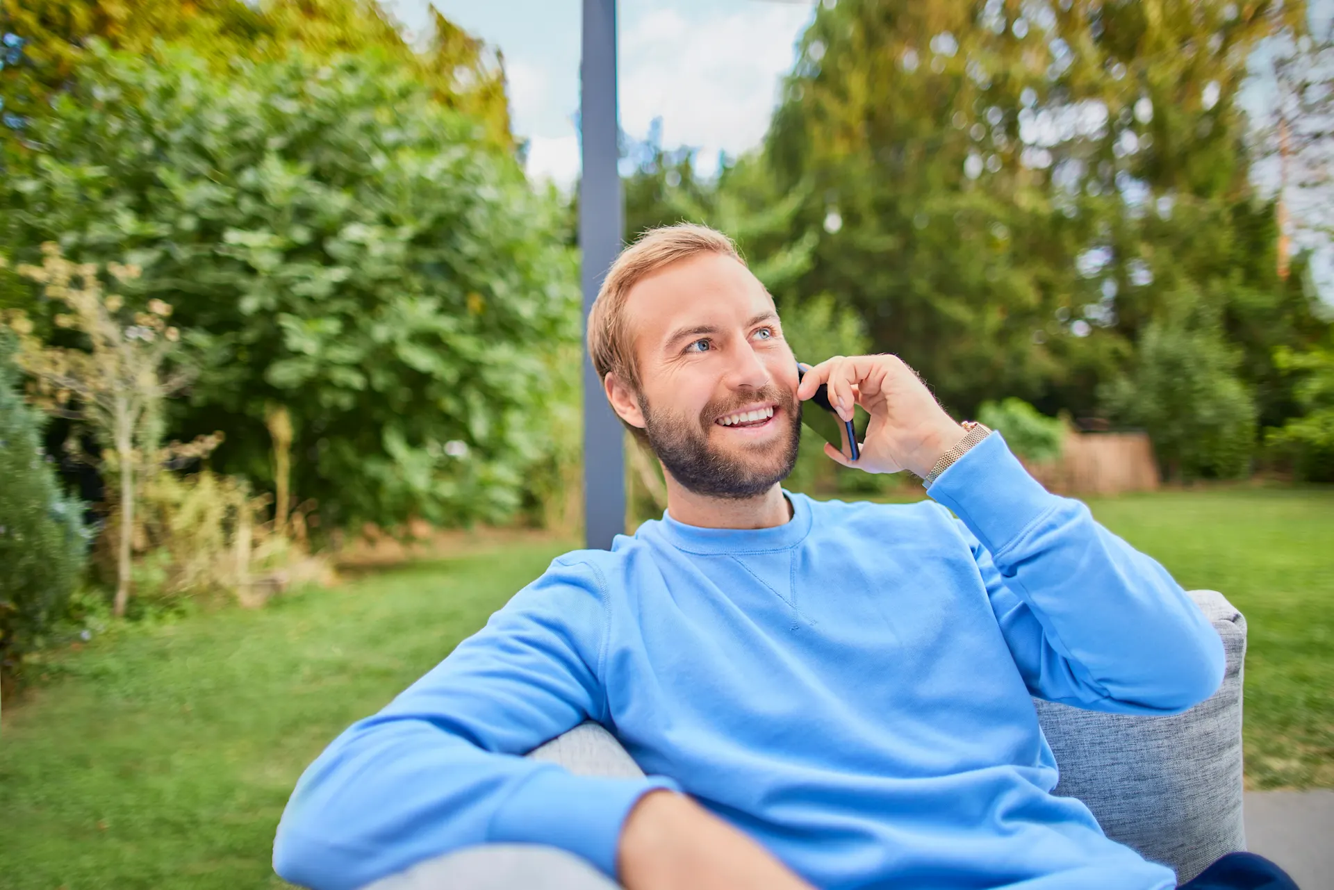 Ein Mann sitzt mit einem Smartphone auf einem Sofa im Garten und nutzt den telefonischen Kundenservice der NEW Energie um Strompreise zu vergleichen.