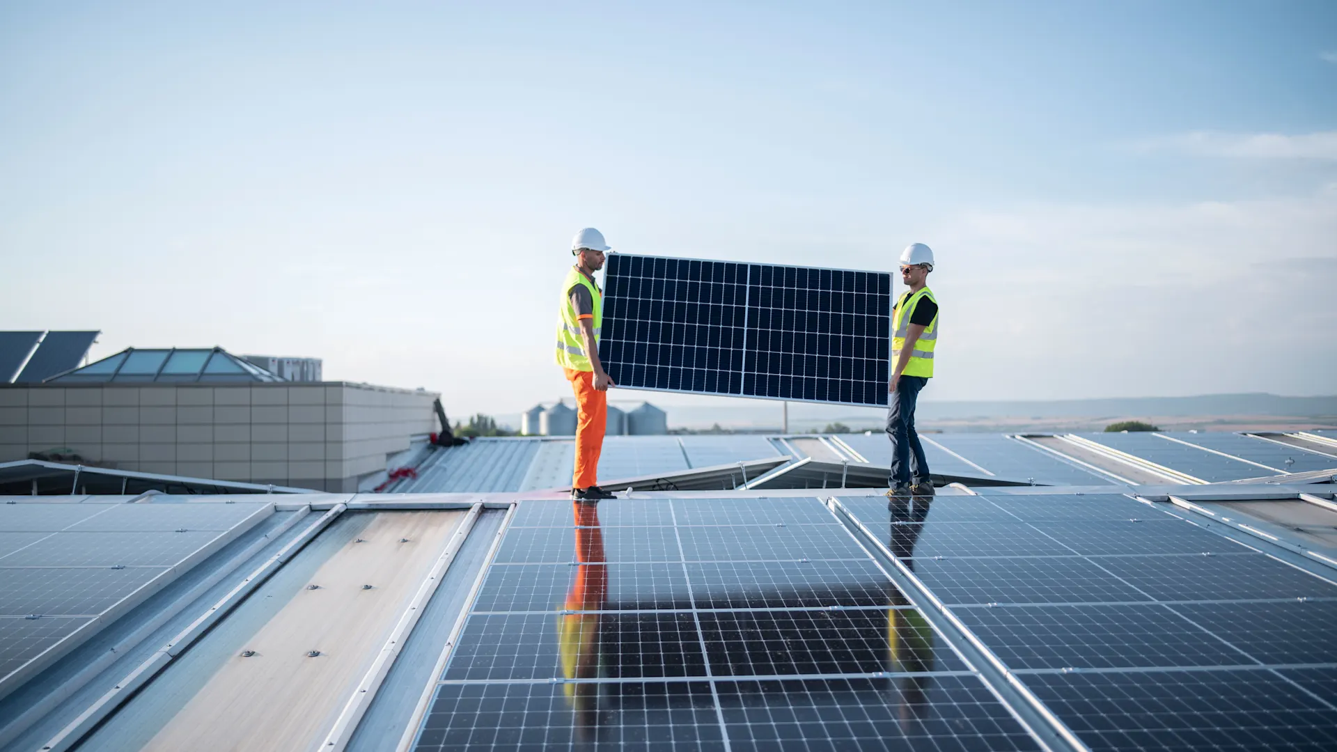 Handwerker mit Photovoltaik Panel in der Hand auf dem Dach Zwei Techniker stehen auf einem Dach und montieren eine PV-Anlage auf Geschäftsräumen.