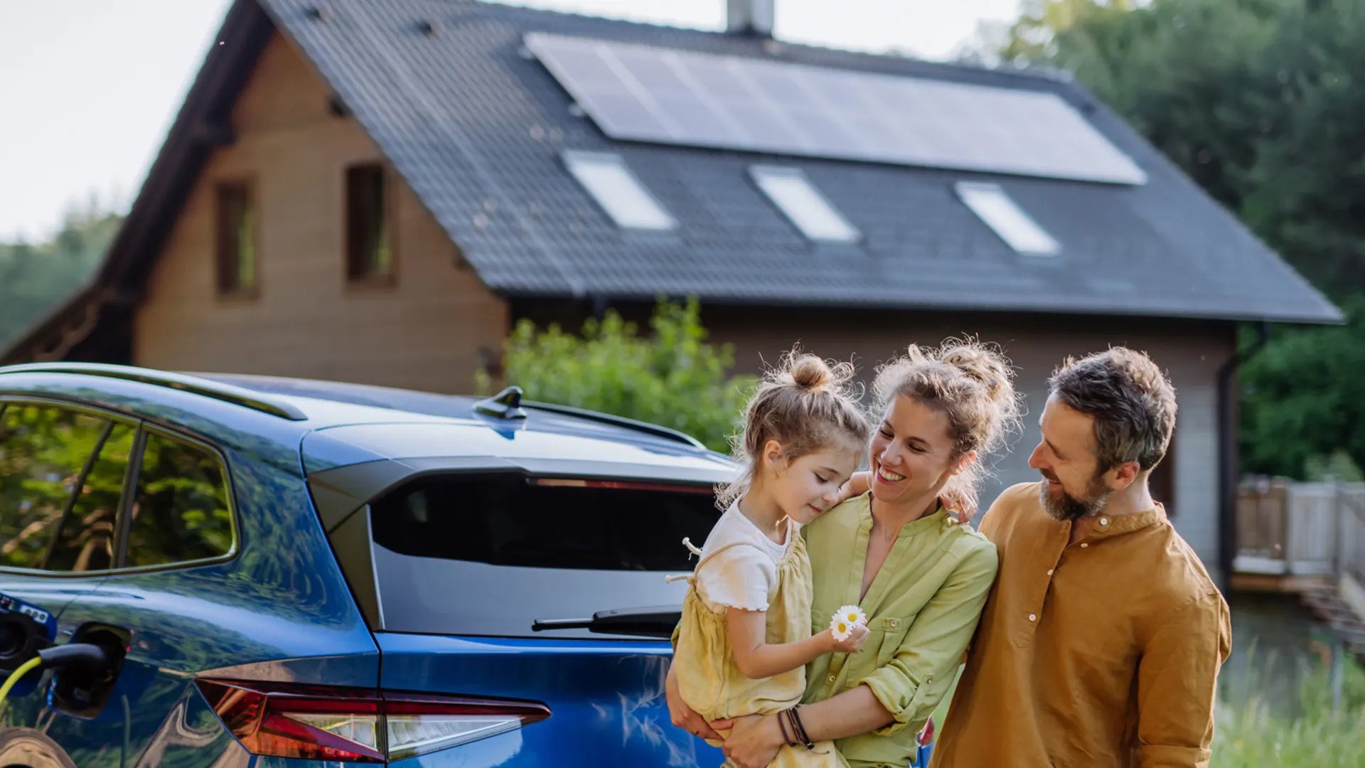 Familie steht vor Ihrem Haus mit einer PV-Anlage auf dem Dach Familie steht vor einem mit Haus im Grünen, mit einer PV-Anlage auf dem Dach und einem Elektroauto davor.