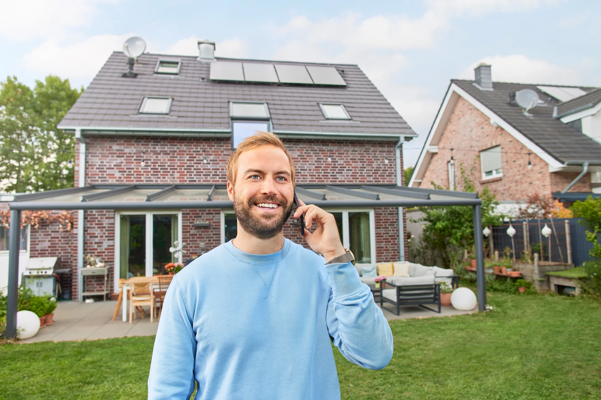 Ein zufriedener Photovoltaikanlagen Besitzer telefoniert vor seinem Einfamilienhaus, auf dessen Dach die Photovoltaikanlage zu sehen ist.