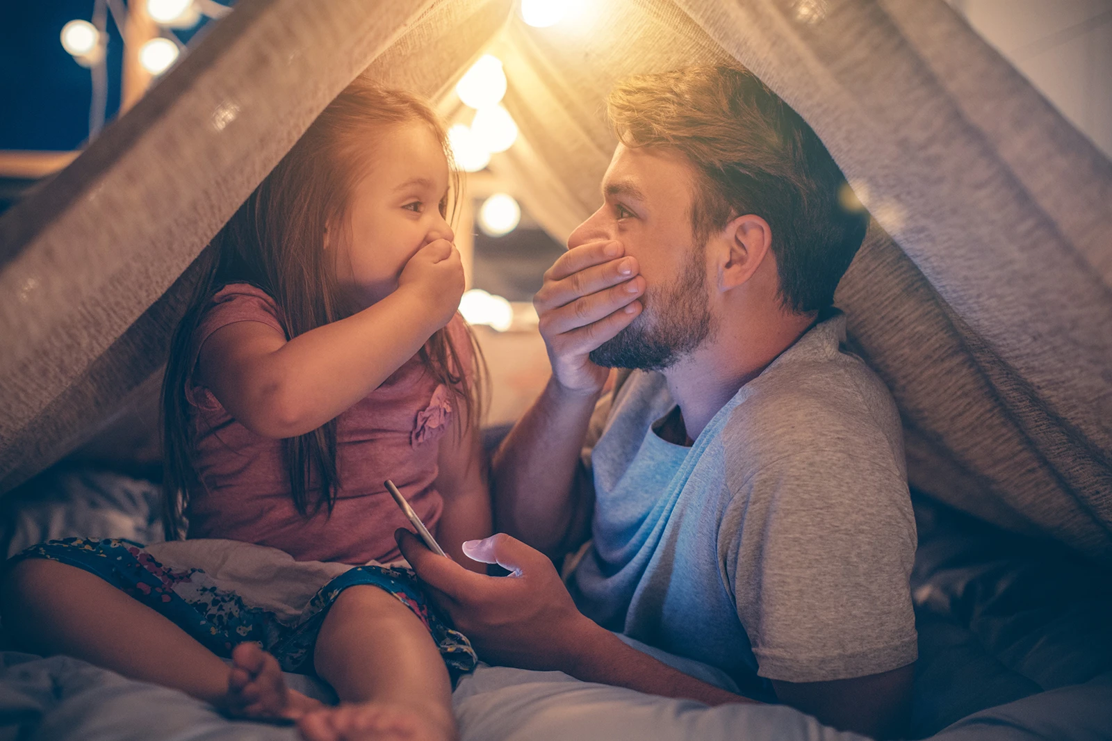 Vater und Tochter sitzen im Wohnzimmer unter einer Deckenhöhle und schauen gemeinsam aufs Smartphone und freuen sich über günstigen Strom.
