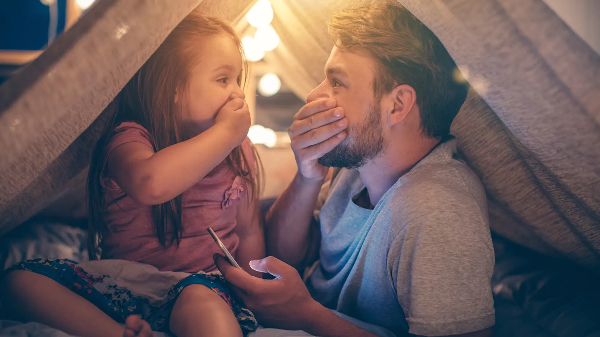 Vater und Tochter sitzen im Wohnzimmer unter einer Deckenhöhle und schauen gemeinsam aufs Smartphone und freuen sich über günstigen Strom.