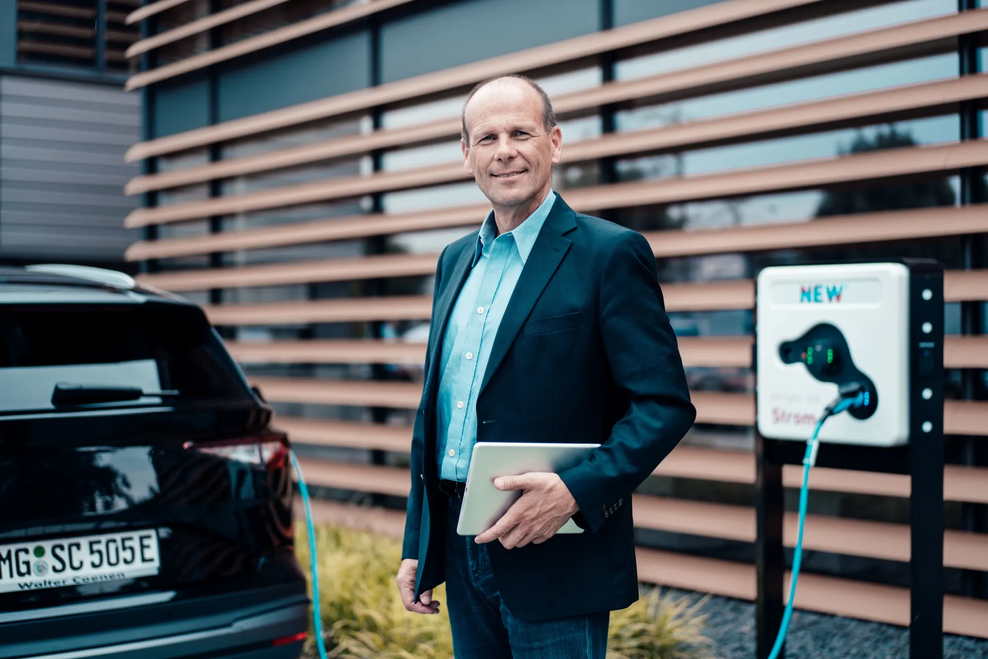 Geschäftsmann steht vor seinem E-Auto mit einem Tablet in der Hand.