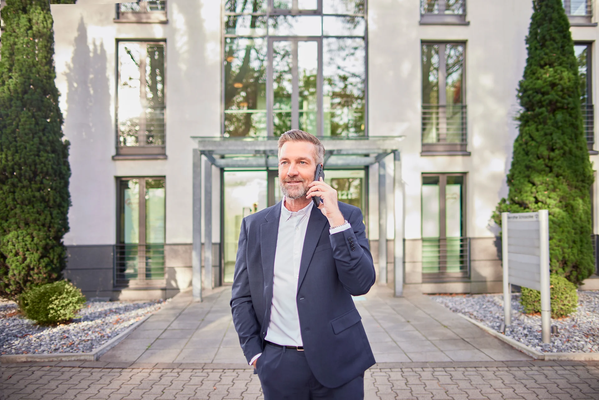 Ein Mann im Anzug steht telefonierend vor einem Mehrfamilienhaus.
