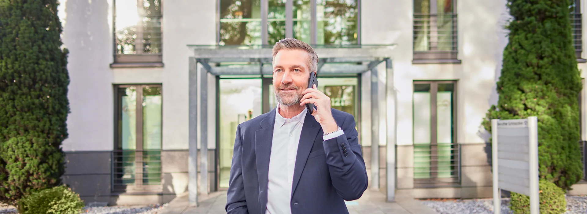 Mann telefoniert mit der NEW Energie Ein Mann im Anzug steht telefonierend mit der NEW Energie vor einem Mehrfamilienhaus.