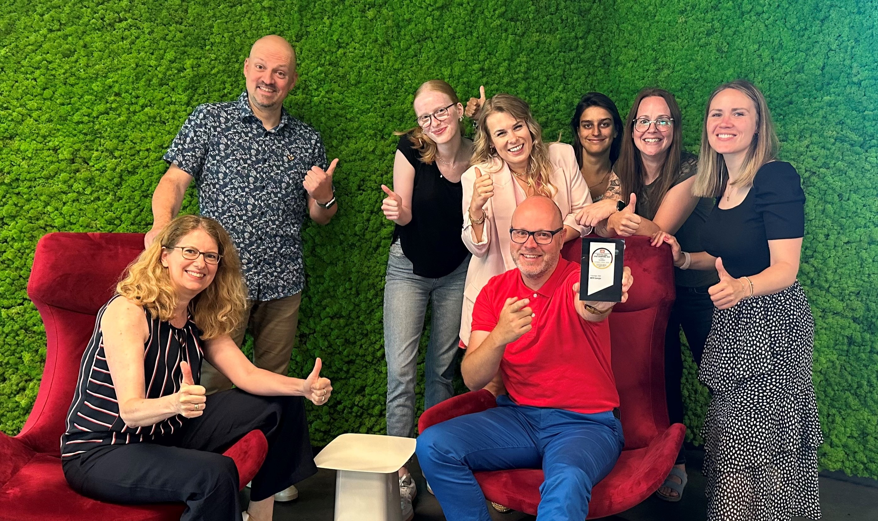 Mitarbeiter:innen der NEW Energie vor einer grünen, blätterreichen Wand. Sie halten die Auszeichnung von Deutschlands Spar-Champions 2025 freudig in den Händen.