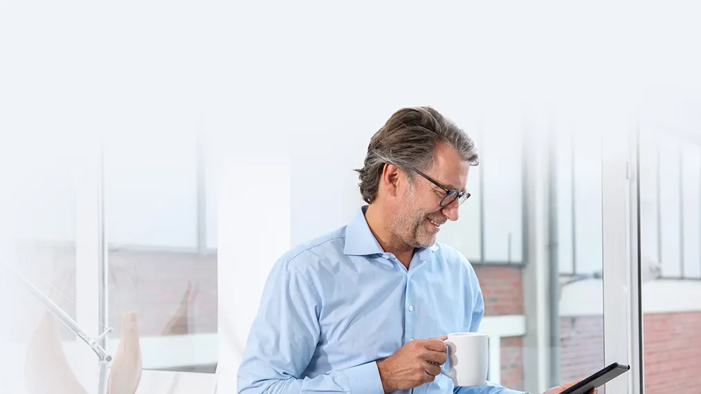 Ein Hausverwalter steht mit einem Kaffee in der Hand vor einem Fenster und schaut mit dem Smartphone nach Serviceangeboten.