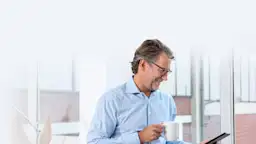 Hausverwalter informiert sich über Serviceangebote der NEW Energie Ein Hausverwalter steht mit einem Kaffee in der Hand vor einem Fenster und schaut mit dem Smartphone nach Serviceangeboten der NEW Energie.